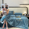 A teal comforter with gold embroidery is displayed on a bed, next to a bedside table and a book.