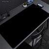A black oversized non-slip desk mat is shown on a gray desk with a laptop, a potted plant, and a small electronic device.