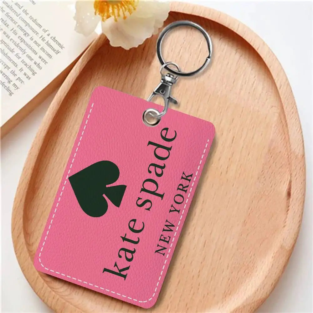 A pink leather card holder with a black spade charm and a metal keyring resting on a wooden board.