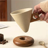 A hand holds a beige ceramic coffee mug with a wooden handle and a matching wooden base and spoon.