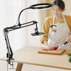 A woman is using an adjustable phone stand with overhead lighting to prepare food on a wooden table.