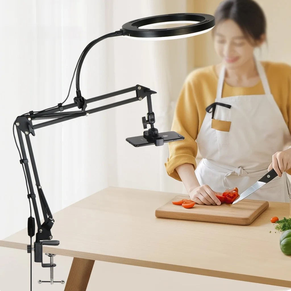 A woman is using an adjustable phone stand with overhead lighting to prepare food on a wooden table.