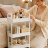 A woman is using a white 3-tier rolling utility cart with wheels for storage in a bright room.