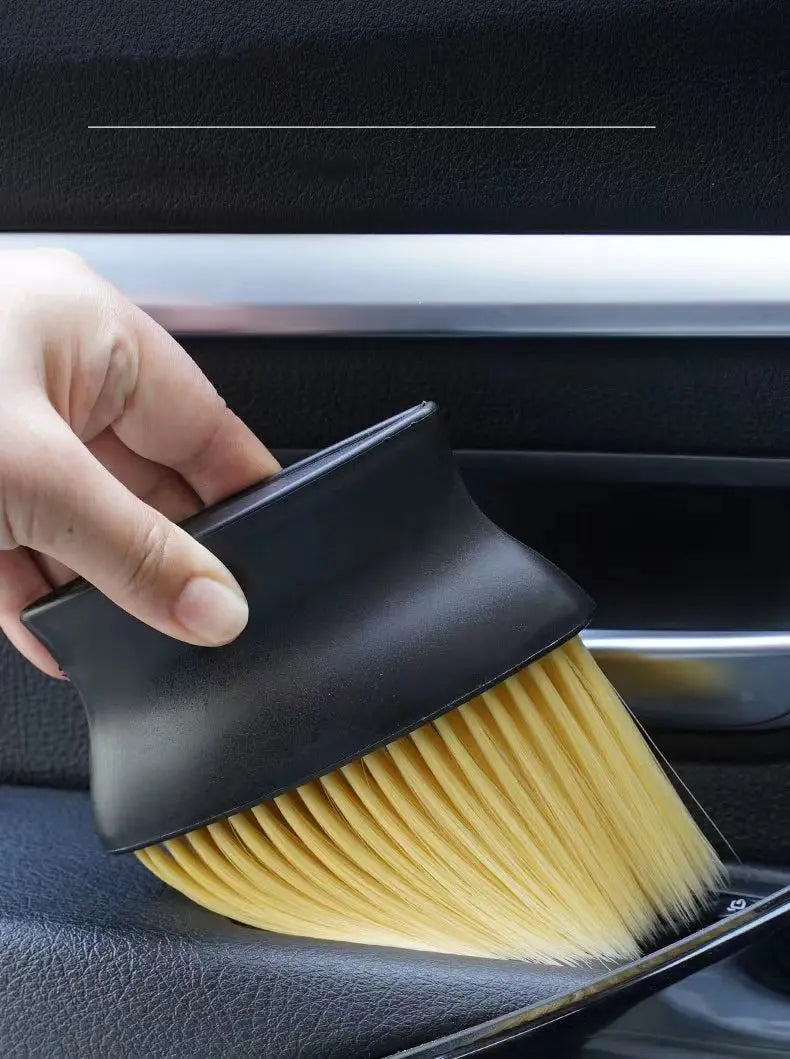 A hand is using a black car interior detailing brush set with soft yellow bristles on a car seat.