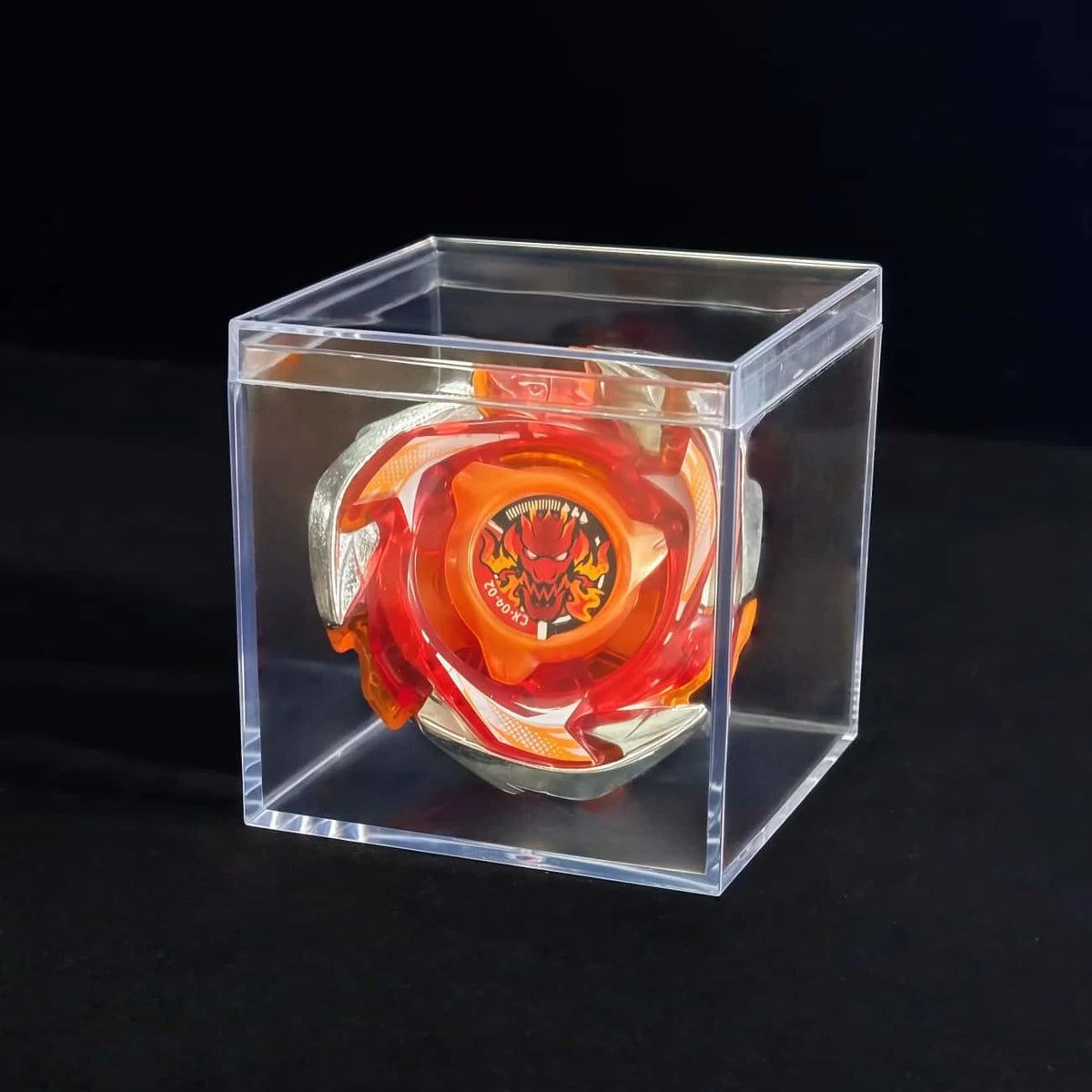 A metal spinning top with a launcher is displayed inside a clear plastic box against a dark background.