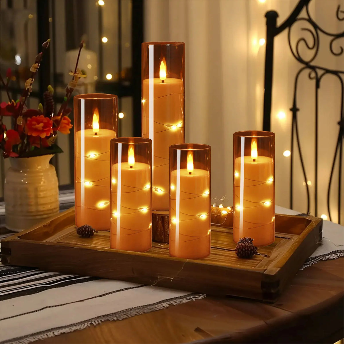 A set of glowing orange LED candles in glass holders displayed on a wooden tray with twinkling string lights in the backgroun