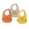 Three silicone baby bibs in peach, beige, and yellow with personalized names
