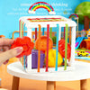 A hand places various colored plastic shapes and balls into a white Montessori sensory toy with colorful hanging strips.