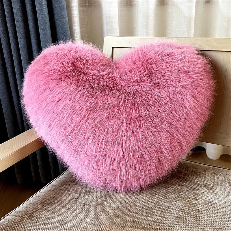 A fluffy pink heart-shaped throw pillow resting on a wooden surface next to a dark curtain.