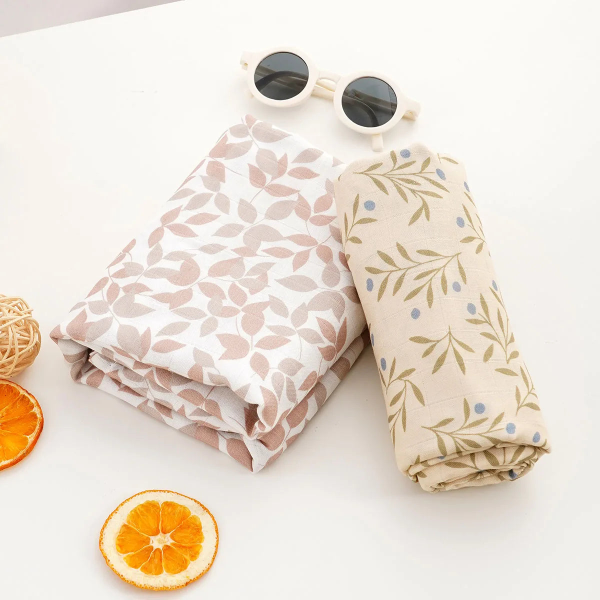 A rolled muslin baby swaddle blanket in beige and green floral print next to sunglasses and an orange slice.