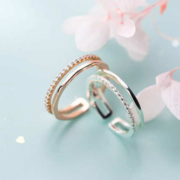 Deux délicats bagues en argent sterling avec des diamants pavés, présentées sur un fond bleu clair avec des fleurs roses.