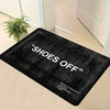 A black minimalist doormat with the text SHOES OFF stamped on it, placed on a light-colored floor.