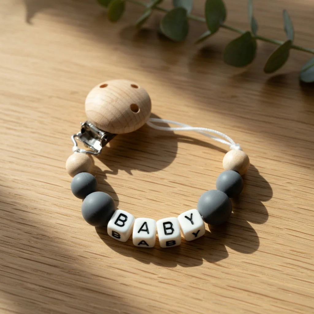 A wooden baby pacifier clip featuring custom name letters and a silicone ring on a light wood background.