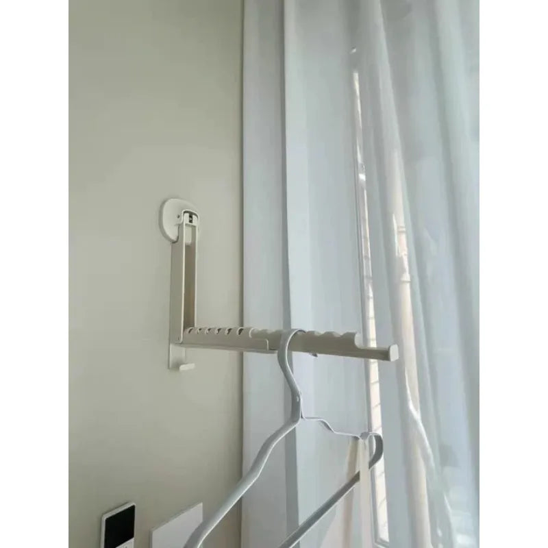 A light beige foldable wall-mounted clothes drying rack with an adhesive mount hanging in front of a sheer white curtain.