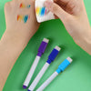 A hand holds a white marker and a piece of paper with colorful paint marks on a green background.