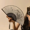 A hand holds a black and white bamboo and silk folding fan with floral designs against a light background.