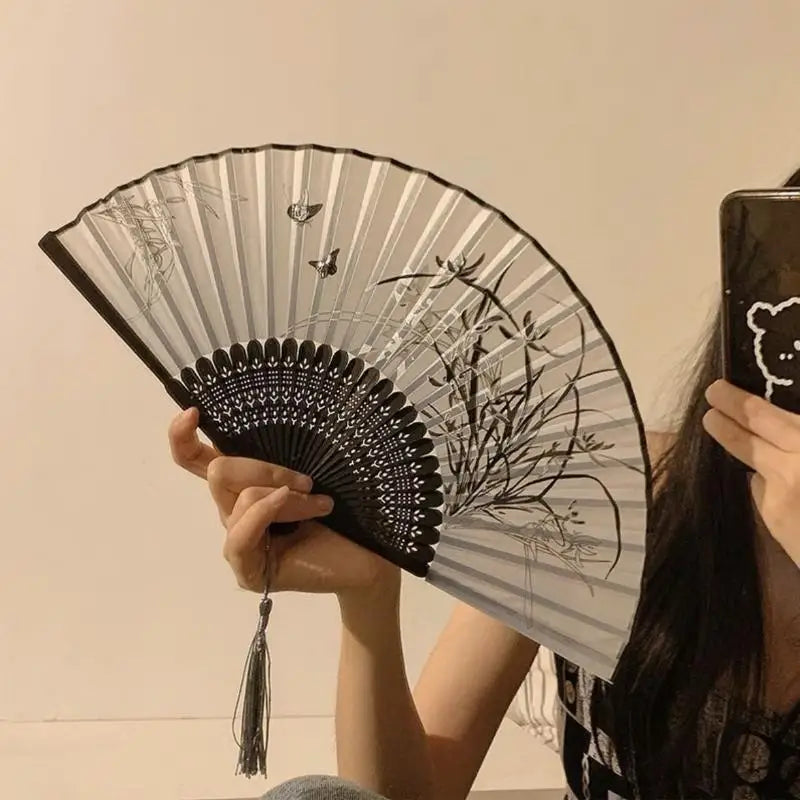 A hand holds a black and white bamboo and silk folding fan with floral designs against a light background.