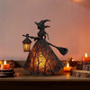 A dark metal witch figure with a broom is displayed inside an orange-toned lantern, illuminating a warm glow.