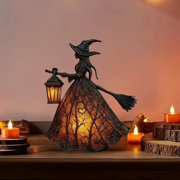 A dark metal witch figure with a broom is displayed inside an orange-toned lantern, illuminating a warm glow.