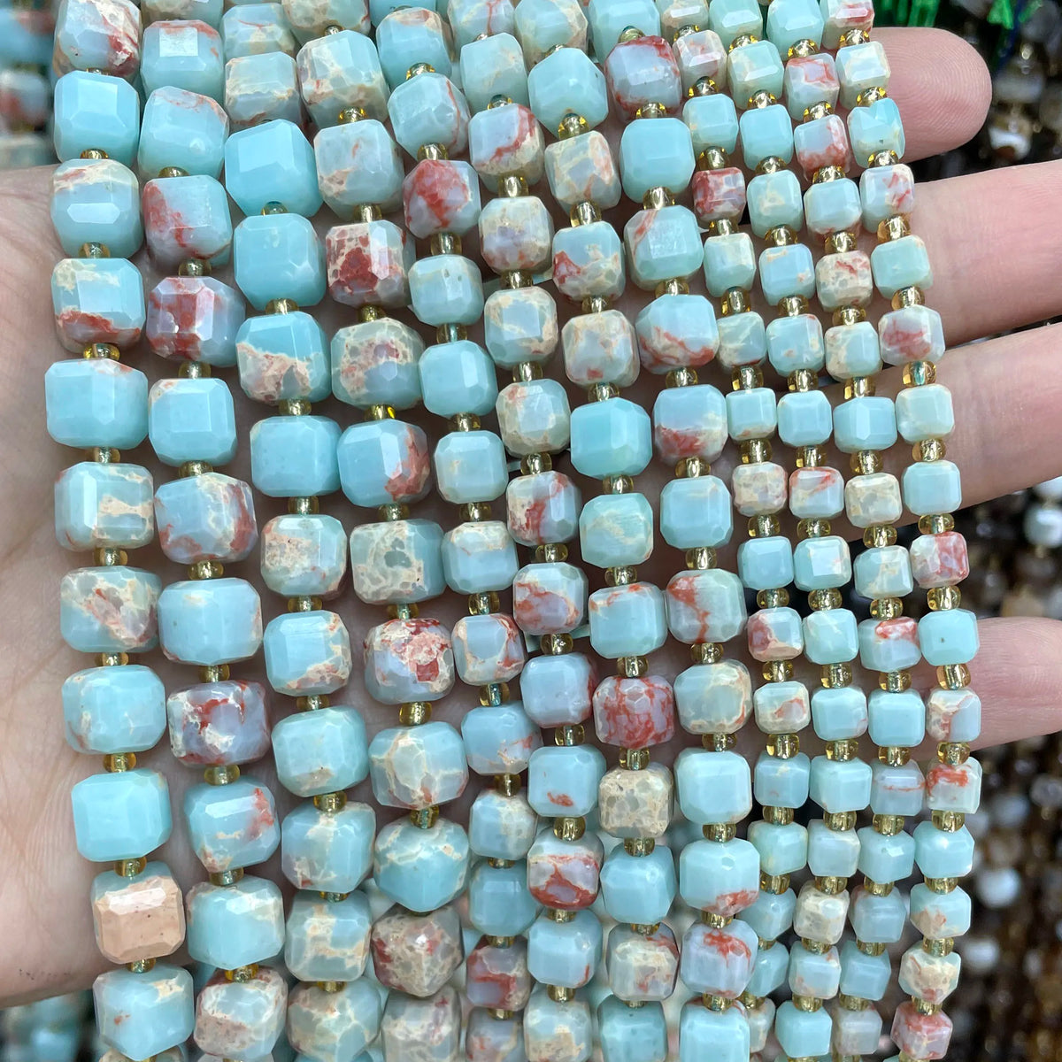 A hand holds a strand of square faceted beads made from natural stone in shades of light blue and pink.
