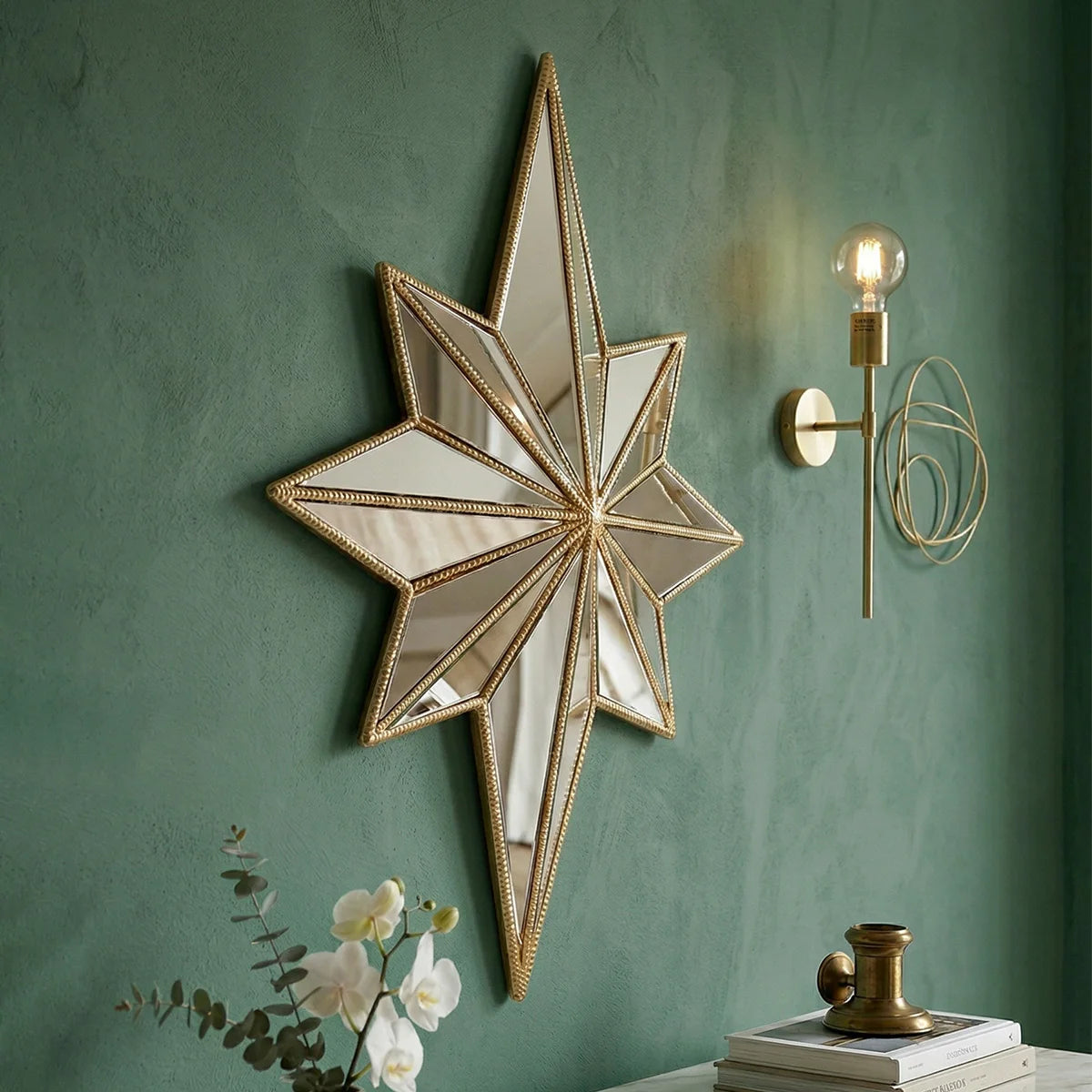 A gold-framed octagonal star resin wall mirror mounted on a green wall with a brass light fixture.