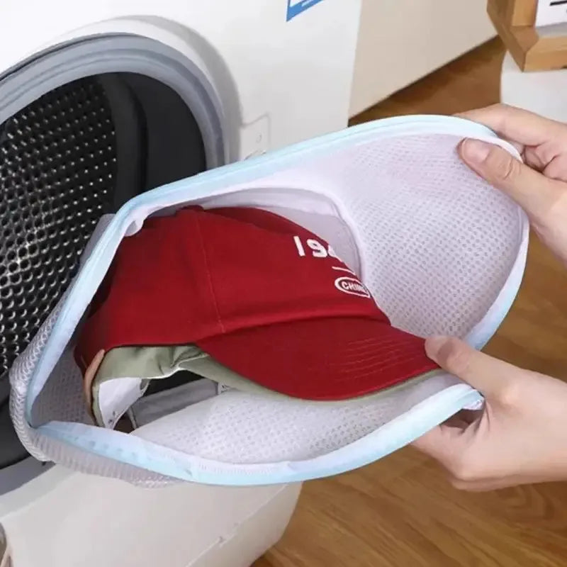 A hand is placing a red baseball cap into a white mesh laundry bag for hats and delicate clothing.