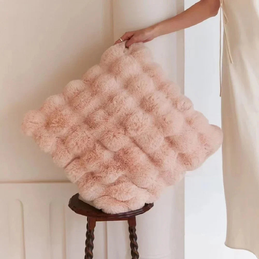A fluffy, light pink decorative pillow cover is being held by a hand against a white wall.