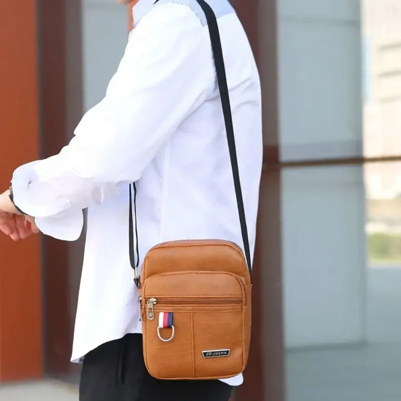 Un homme tient un sac bandoulière en cuir fauve avec une bretelle noire et une étiquette rayée.