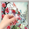 A hand is applying floral stained glass window film to a glass surface, showing pink and red flowers and branches.
