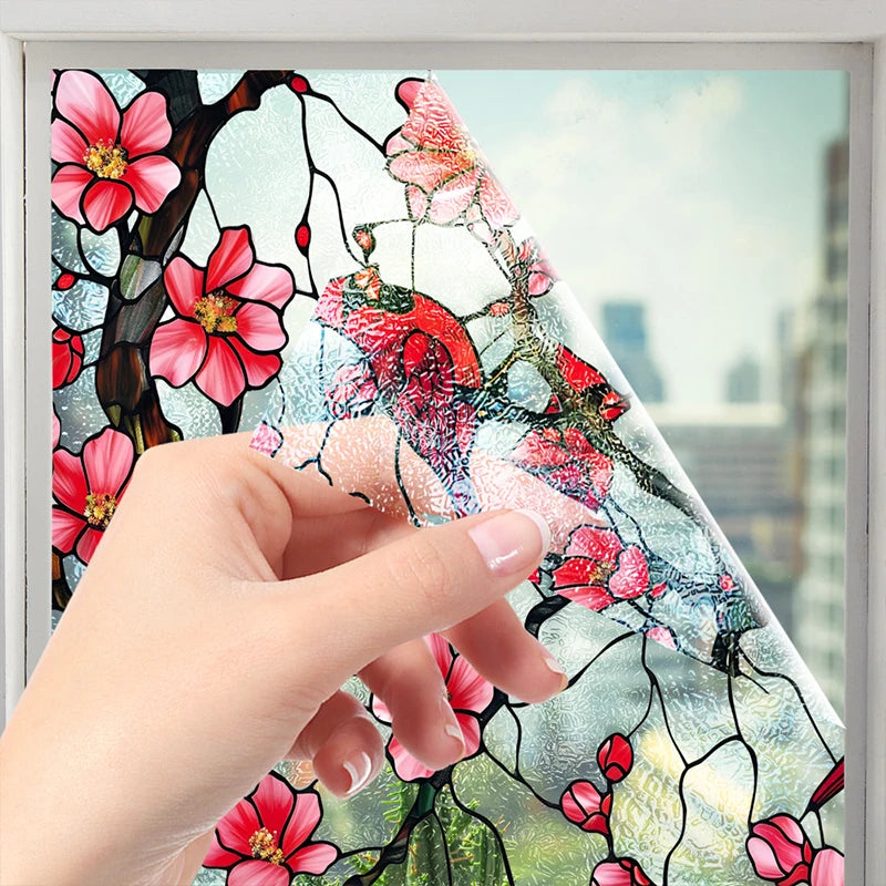 A hand is applying floral stained glass window film to a glass surface, showing pink and red flowers and branches.