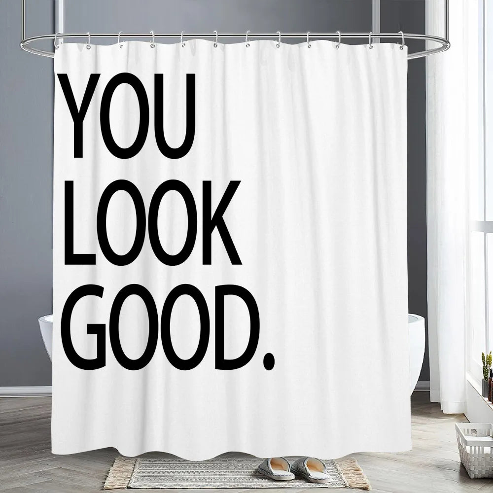A white shower curtain with the text YOU LOOK GOOD in black bold letters hanging from a metal rod.