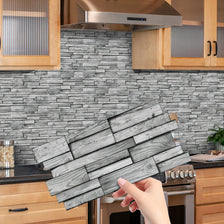 A hand holds a rectangular piece of peel-and-stick tile stickers designed for kitchen and bathroom backsplashes.