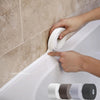 A hand is applying self-adhesive waterproof sealing tape to a gap between a white bathtub and a tiled wall.