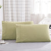 A set of two light olive green microfiber pillowcases resting on a gray bed, near a window with white checkered curtains.