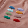 A set of eight oval solid color scarf pins in shades of blue, green, yellow, and pink resting on a peach-colored fabric.