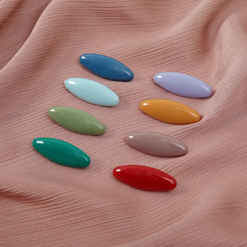 A set of eight oval solid color scarf pins in shades of blue, green, yellow, and pink resting on a peach-colored fabric.