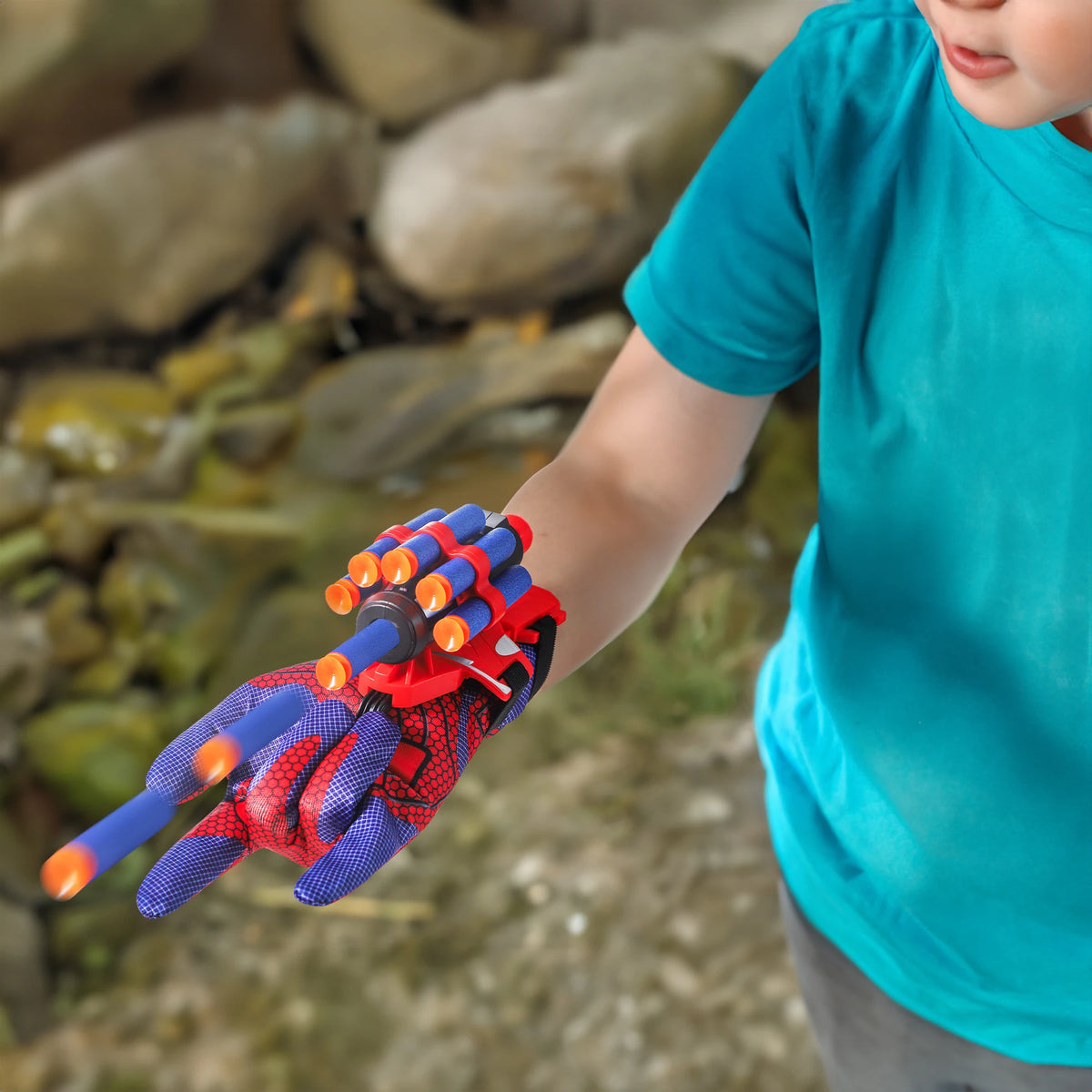 A child is holding a red and blue spider web shooter glove with foam darts in an outdoor setting.