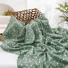 A draped, sage green muslin baby swaddle blanket with a subtle floral pattern is displayed on a white surface.