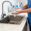 A person in blue scrubs dispenses soap into a stainless steel sink using a faucet, with a black dispenser nearby.