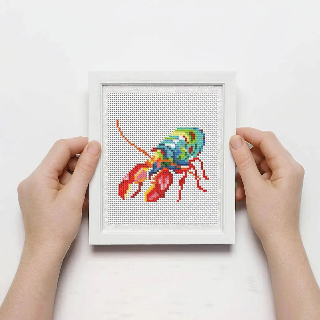 A hand holds a white framed cross stitch kit featuring a colorful sea life design.