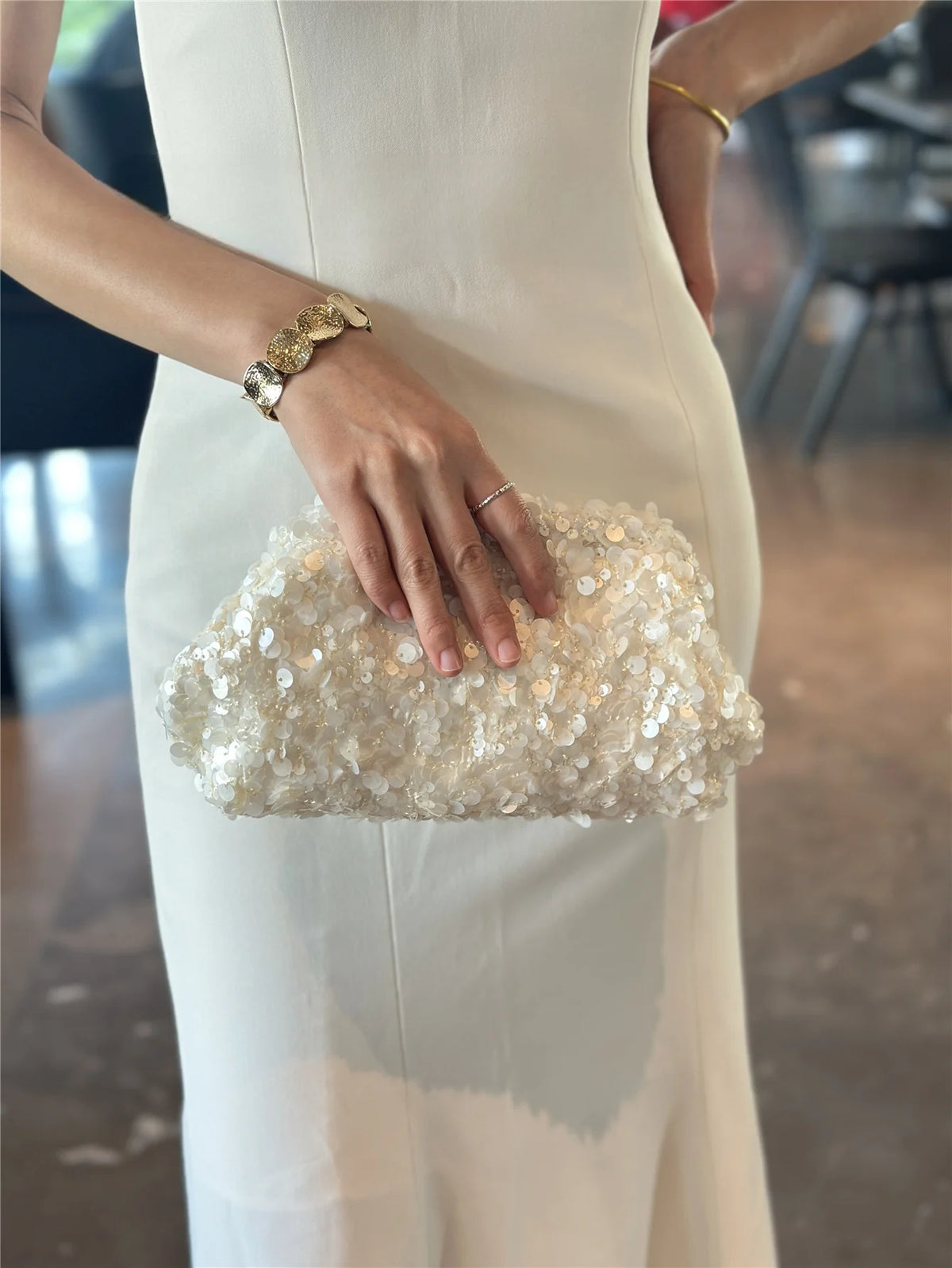 A woman wearing a white dress holds a light gold sequin beaded chain shoulder clutch bag.