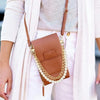 A brown leather purse with a pearl and gold chain is worn by a person in a light pink cardigan.