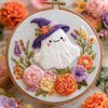 A cross stitch kit featuring a white witch hat and face surrounded by colorful floral embroidery stitches.