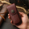 A hand holds a dark reddish-brown genuine leather folding knife case with a decorative metal emblem.