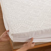 Hands are folding a white waterproof quilted mattress protector with extra padding on a wooden surface.