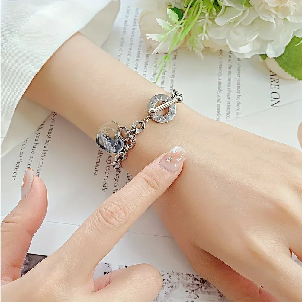 A close-up of a stainless steel double heart pendant bracelet worn on a wrist, resting on a piece of paper with flowers in th