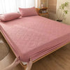 A pink quilted mattress protector is shown on a bed, with a hand adjusting the fabric.