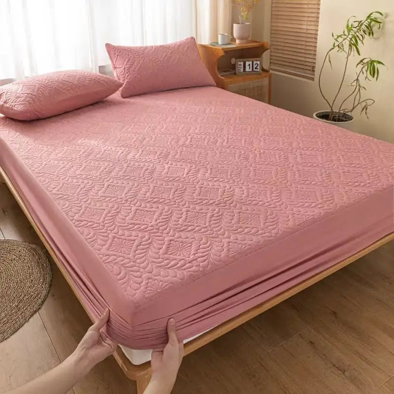 A pink quilted mattress protector is shown on a bed, with a hand adjusting the fabric.