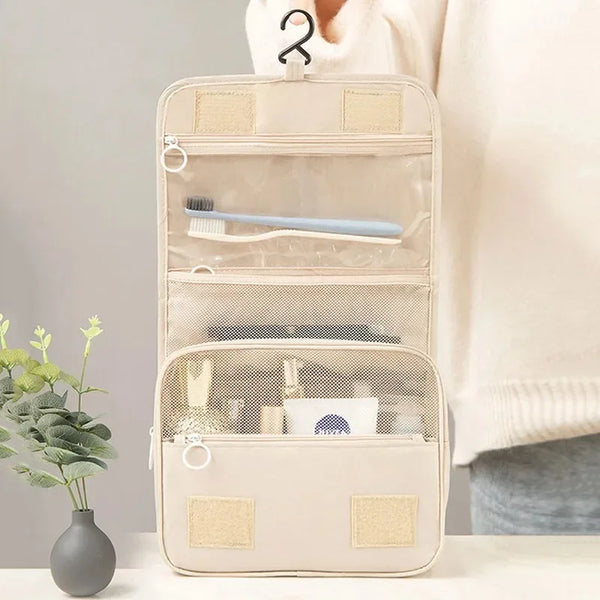 A beige waterproof hanging toiletry bag with clear compartments and a brush inside, held by a person's hand.