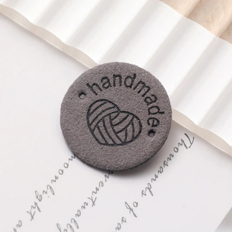 A round, gray PU leather label with an engraved heart design is placed on a white paper background.